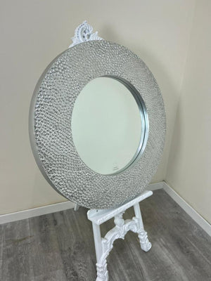 Decorative round mirror with a textured frame on a white stand against a beige wall.