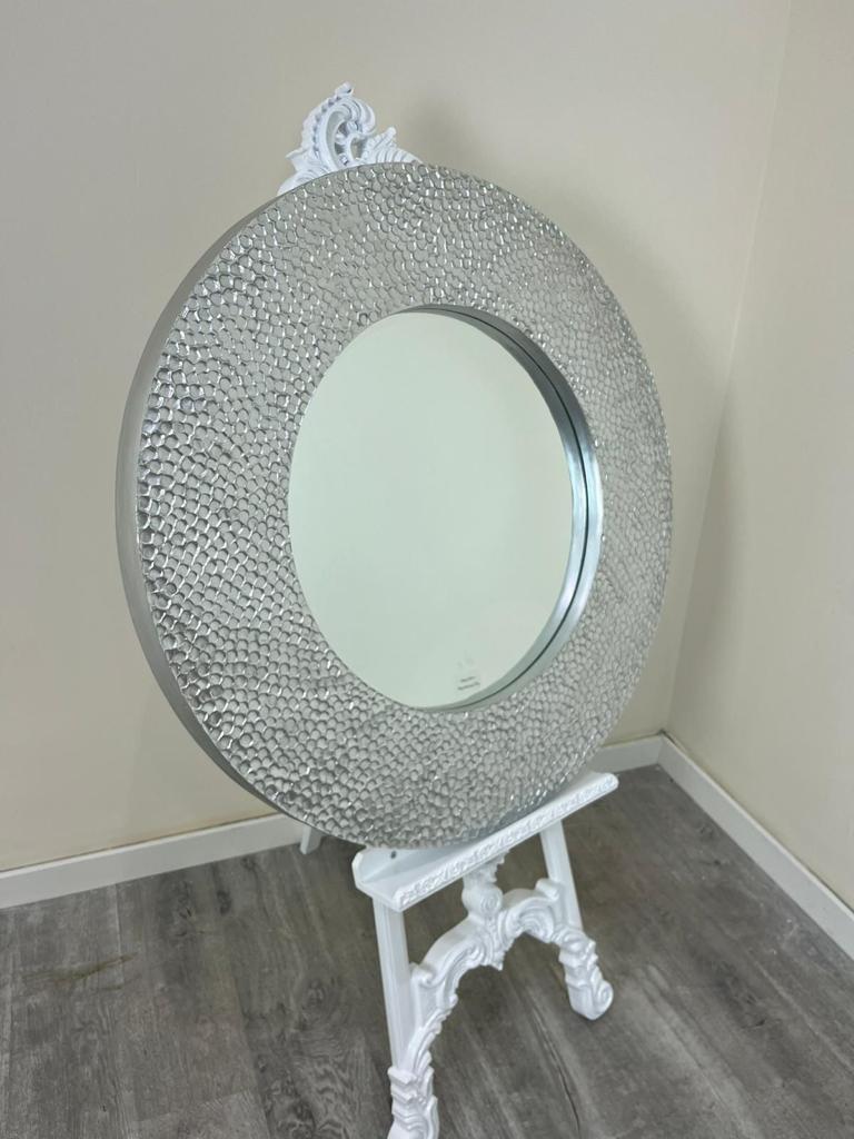 Decorative round mirror with a textured frame on a white stand against a beige wall.