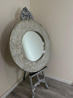 Decorative round mirror with intricate design on a stand against a plain wall.