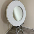 Decorative round mirror with intricate silver frame on a stand against a beige wall.