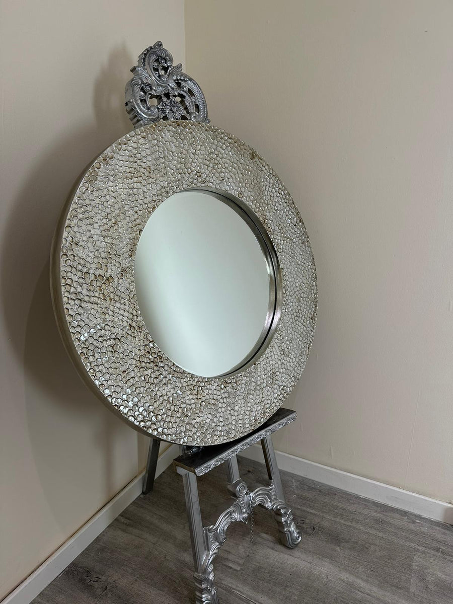Decorative round mirror with intricate design on a stand against a plain wall.