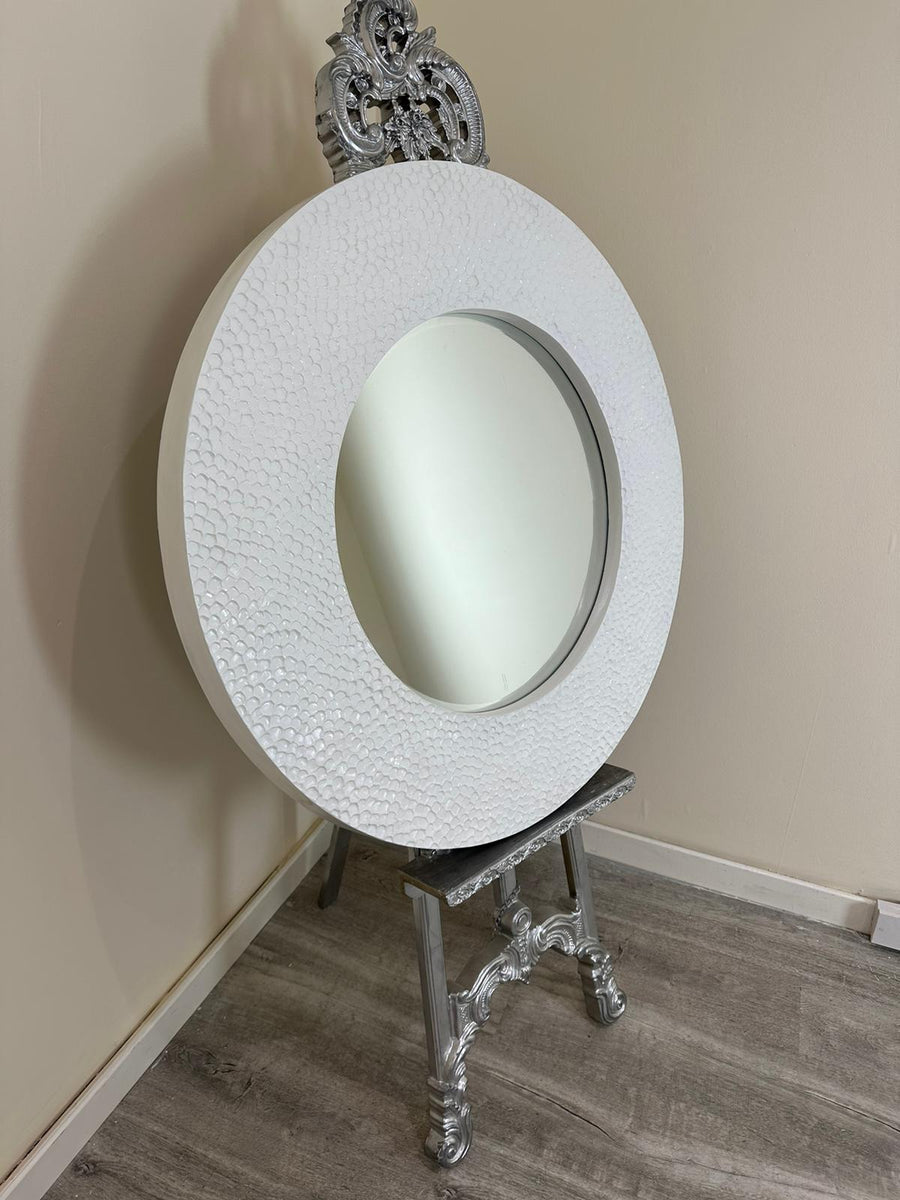 Decorative round mirror with intricate silver frame on a stand against a beige wall.
